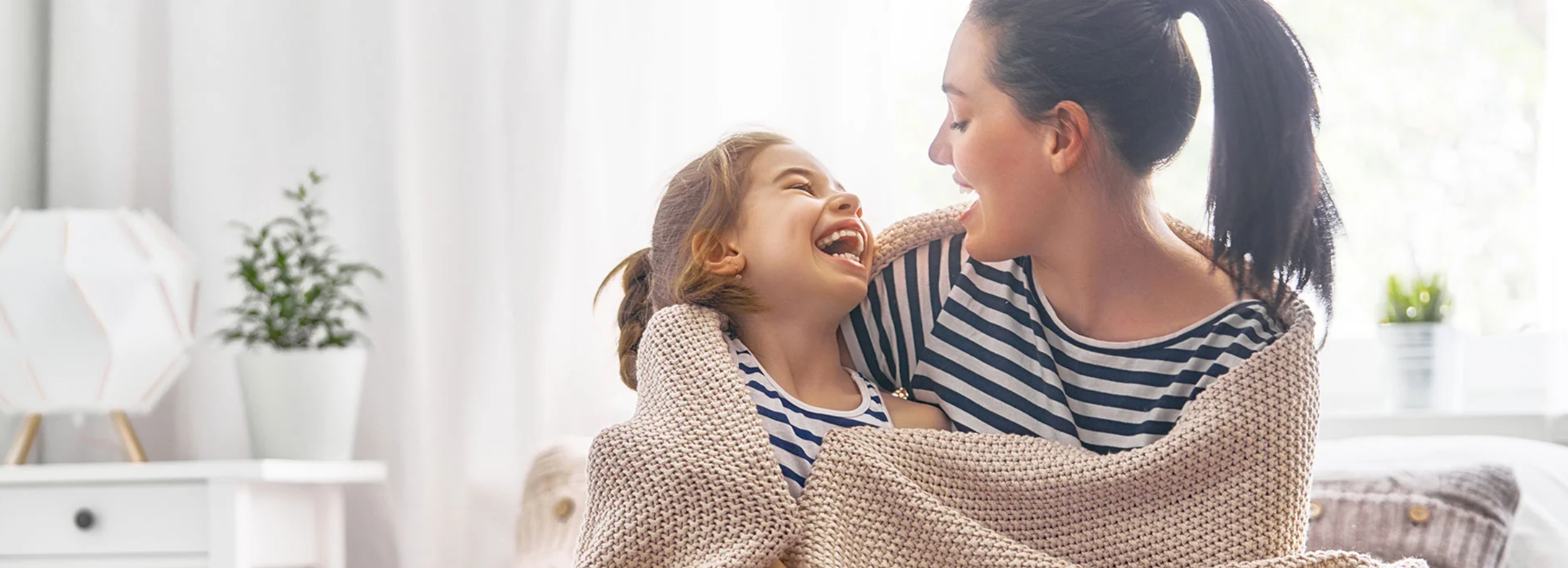 Mother and daughter  wrapped in taupe woven blanket smiling and laughing in brightly lit interior.