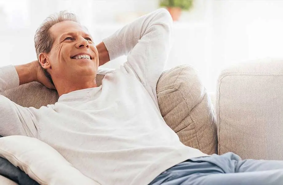 Middle-aged Caucasian man in white sweatshirt smiling and reclining on couch in bright interior