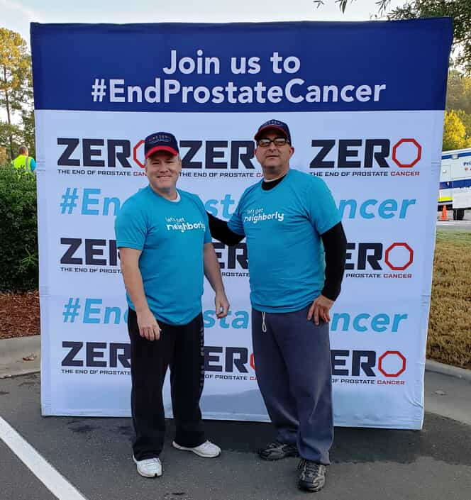 Two Men in teal shirts standing in front of a Zero Prostate Cancer banner