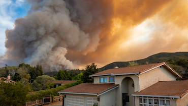 Two story house nestled among hills with large cloud of smoke from wildfire in background.