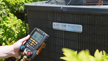 Close-up of digital manifold gauge help up beside Aire Serv branded outdoor HVAC unit.