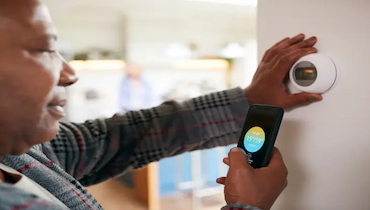 Middle-aged black man using app on smartphone to adjust smart thermostat hanging on wall.