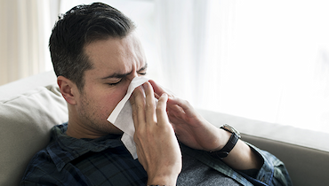 Man blowing nose into tissue on couch in brightly interior.