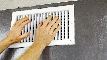 A pair of adult male hands feeling the flow of air coming out of an air vent on a wall near a ceiling in a residential home.